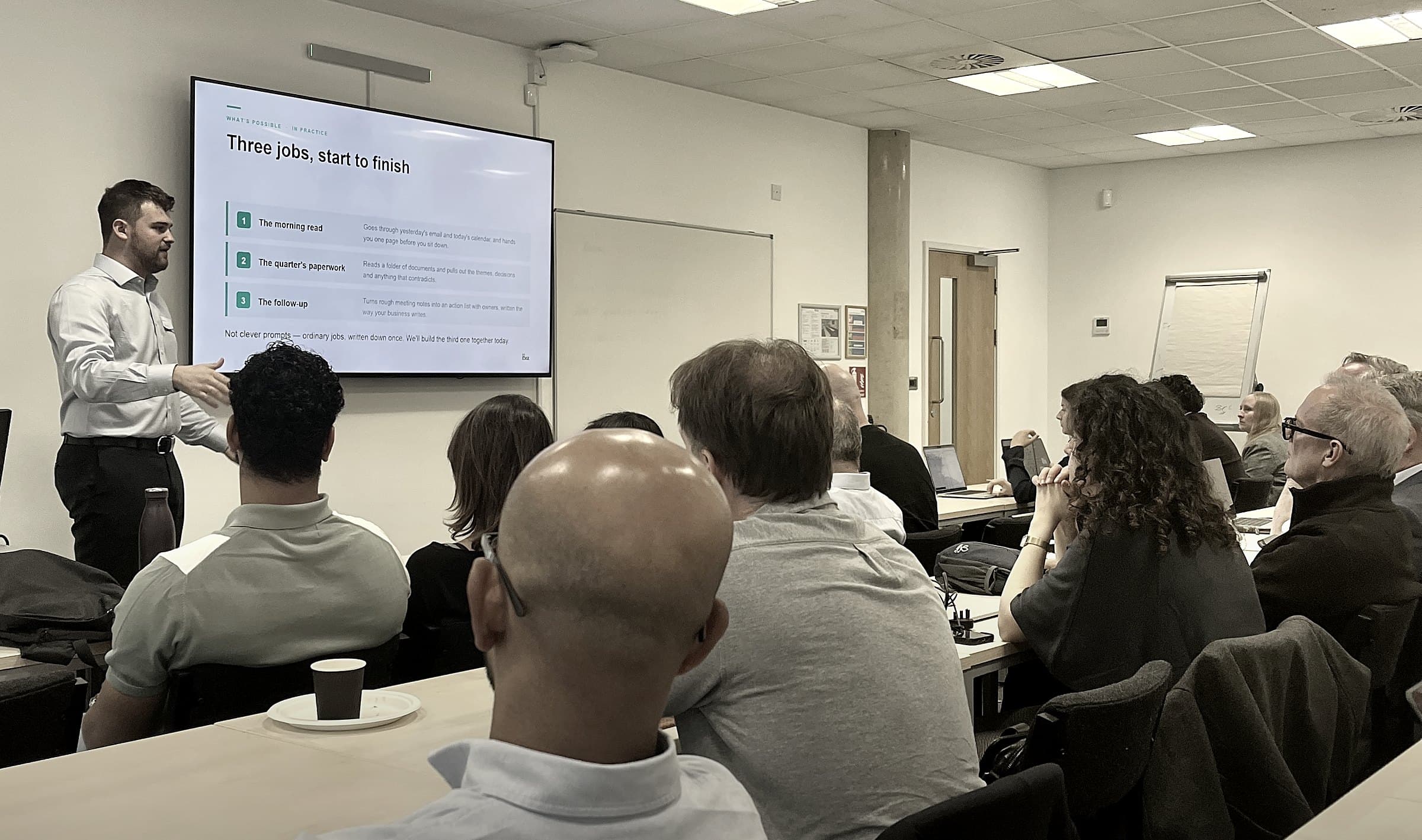 A full room of business owners watching an AI Edge session at the Thames Valley AI Hub in Reading, with a slide on screen showing three everyday jobs being worked start to finish